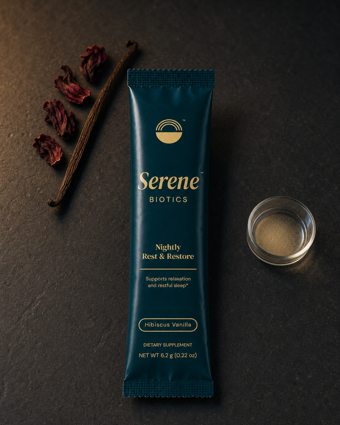 A Nightly Rest & Restore Hibiscus Vanilla stick pack on dark stone in lamplight, beside dried hibiscus petals, a vanilla pod, and a small glass dish of the powder