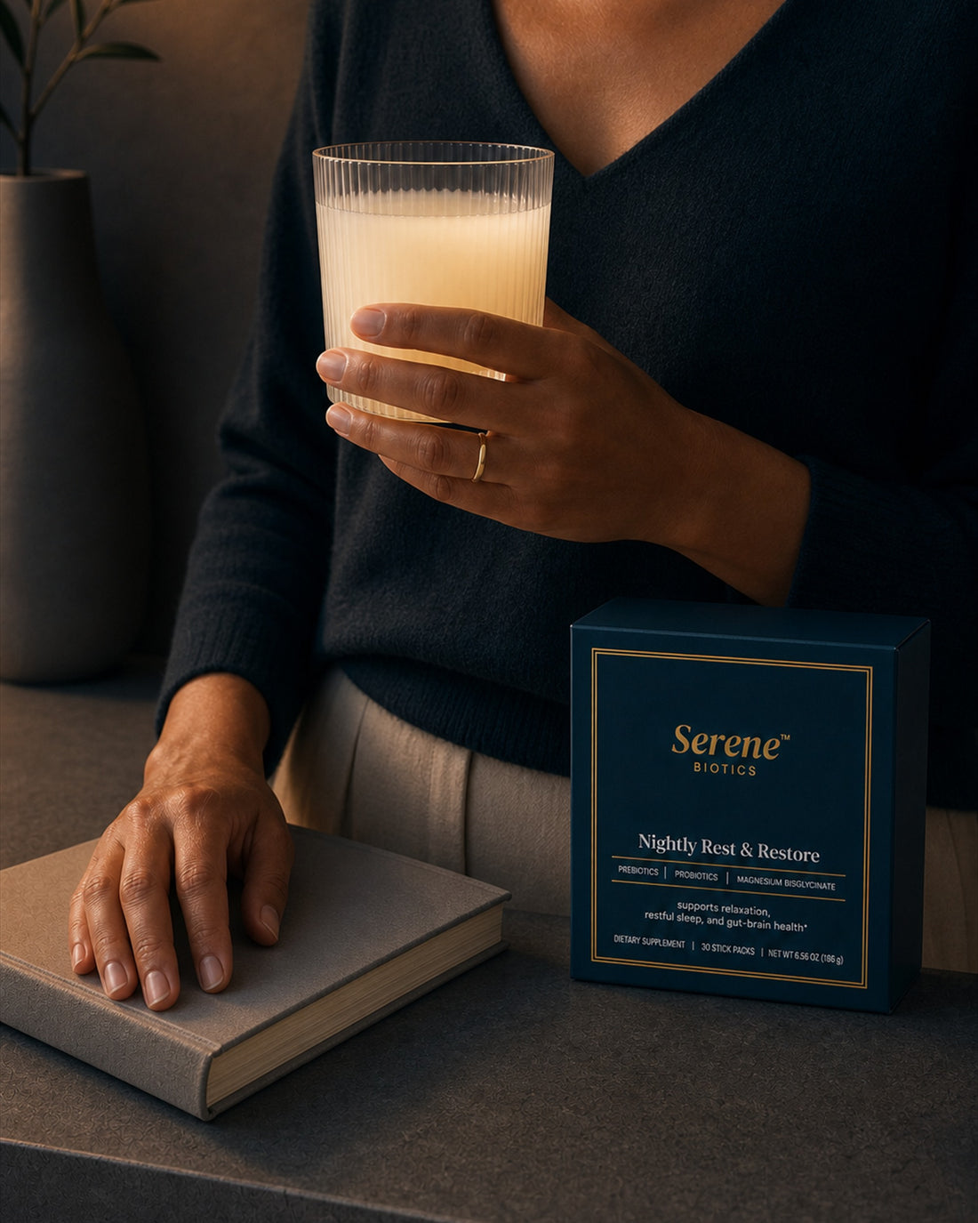 The evening: the prepared pale drink held in a ribbed glass under lamplight, navy knit, the Nightly Rest & Restore carton standing on the counter